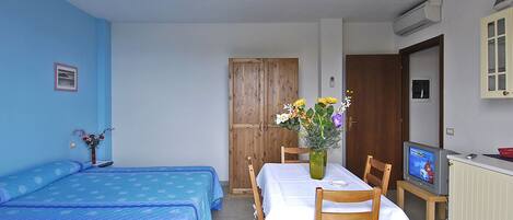 A room with two beds, a dining table with chairs, a vase with flowers, a television, and an air conditioning unit.
