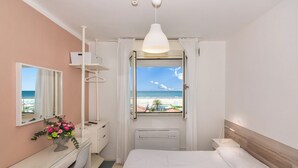 A bedroom with a bed, a desk, a dresser, a mirror, and a window with a beach view. - Happy Hotel (Pietrasanta)