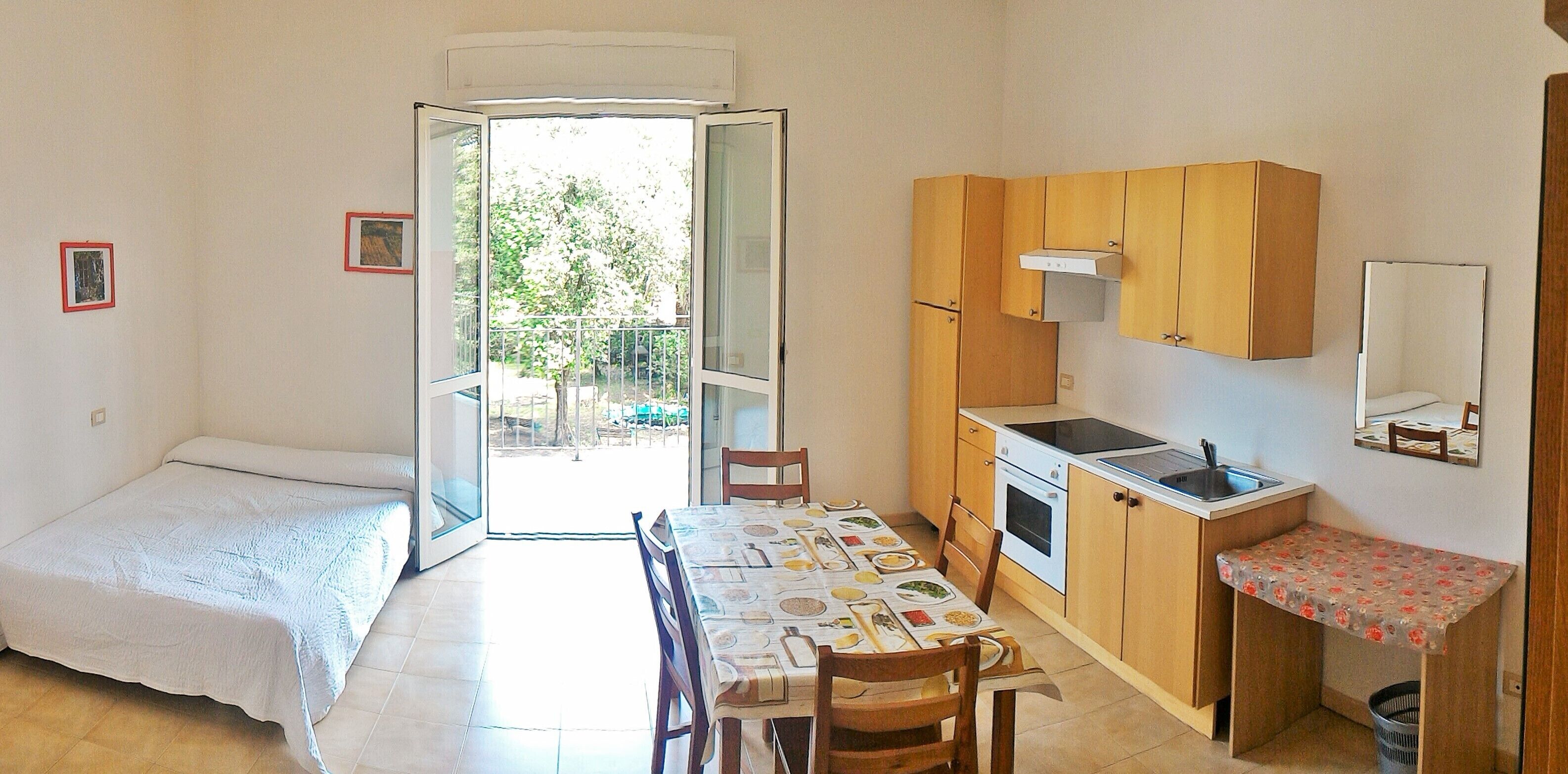 A compact room with a bed, a dining table with chairs, a kitchenette, and a sliding glass door leading to an outdoor area.