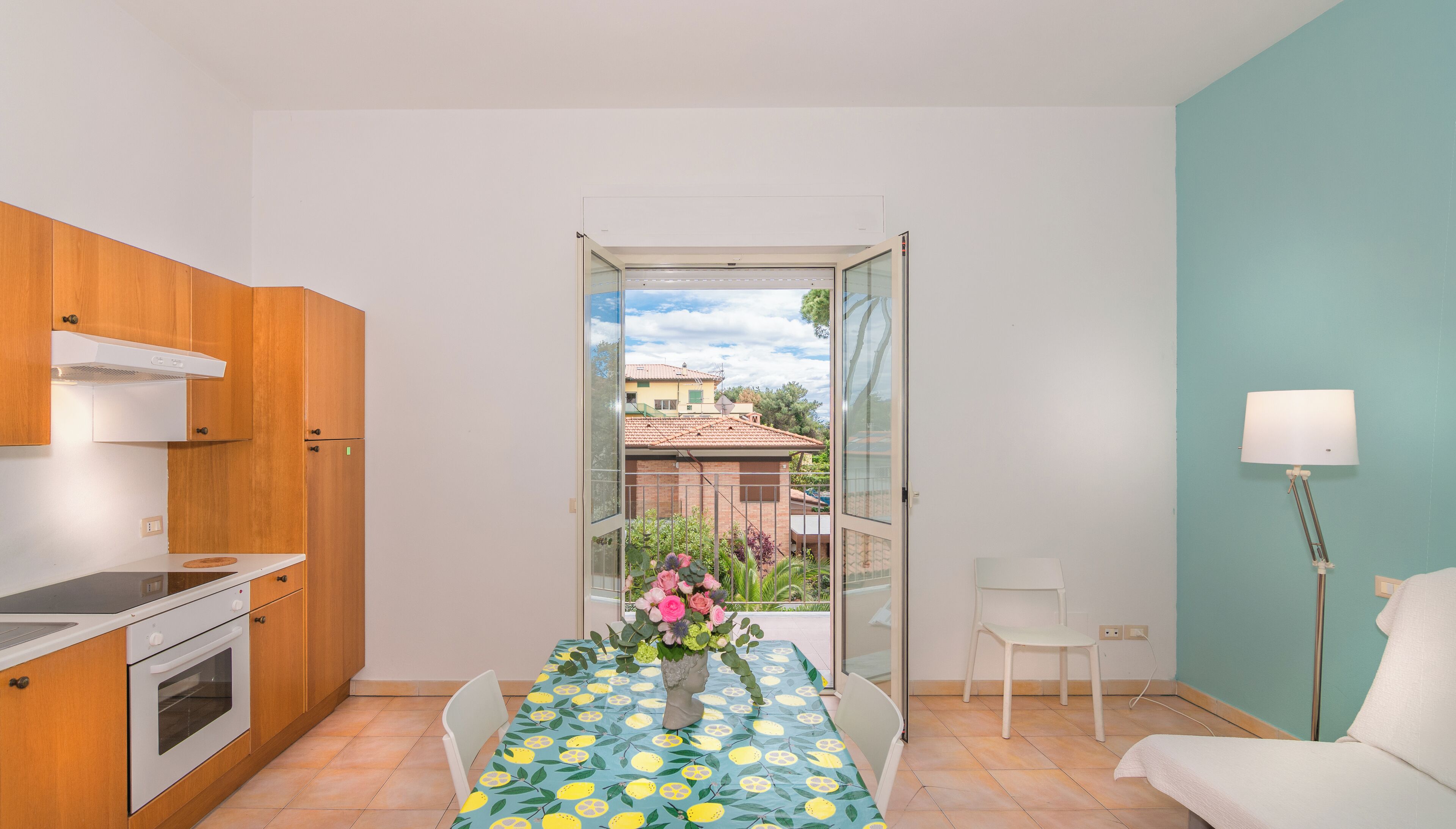 Una cocina funcional con armarios de madera, una estufa blanca y un comedor con una mesa preparada para dos personas. Se ve un balcón con vistas a un edificio y vegetación a través de una puerta abierta.