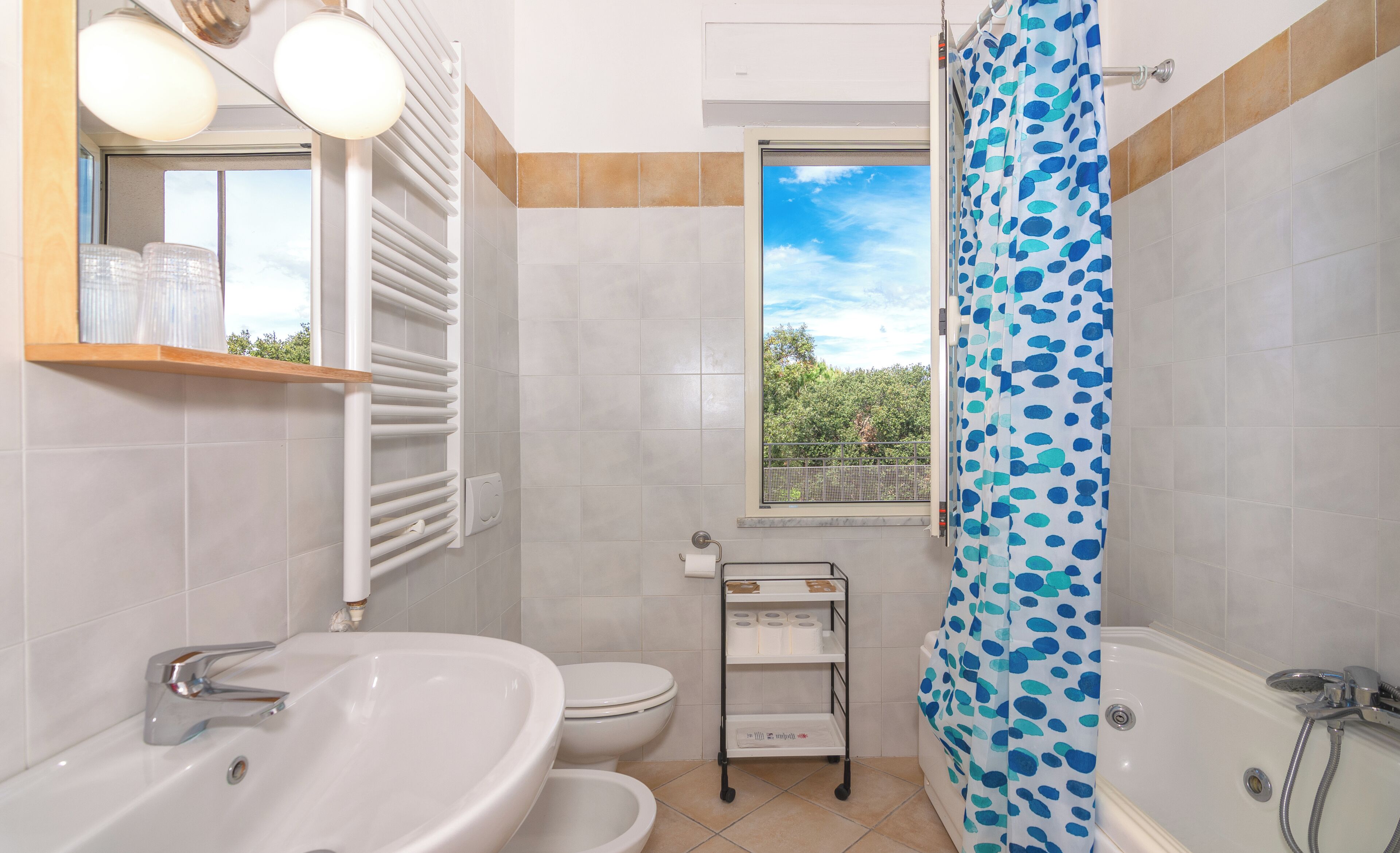 Ein Badezimmer mit Badewanne, Waschbecken, Toilette, einem Fenster mit Blick auf Grünflächen und einem Duschvorhang mit Punkte-Muster.
