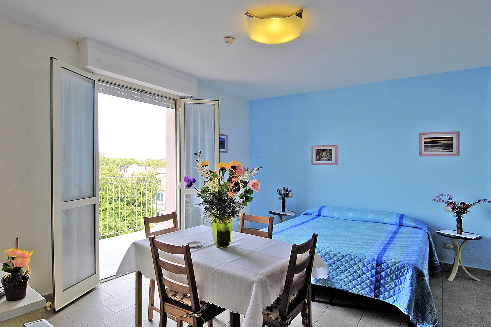 A room with a bed, a table with chairs, a vase with flowers, and a balcony with a railing.