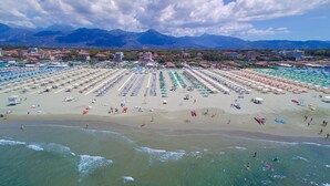 A beach with rows of umbrellas and beach chairs, a clear view of the ocean, and mountains in the background. - Happy Hotel (Pietrasanta)