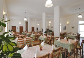A spacious dining hall with multiple tables and chairs, each with a tablecloth and a flower centerpiece. - Happy Hotel (Pietrasanta)