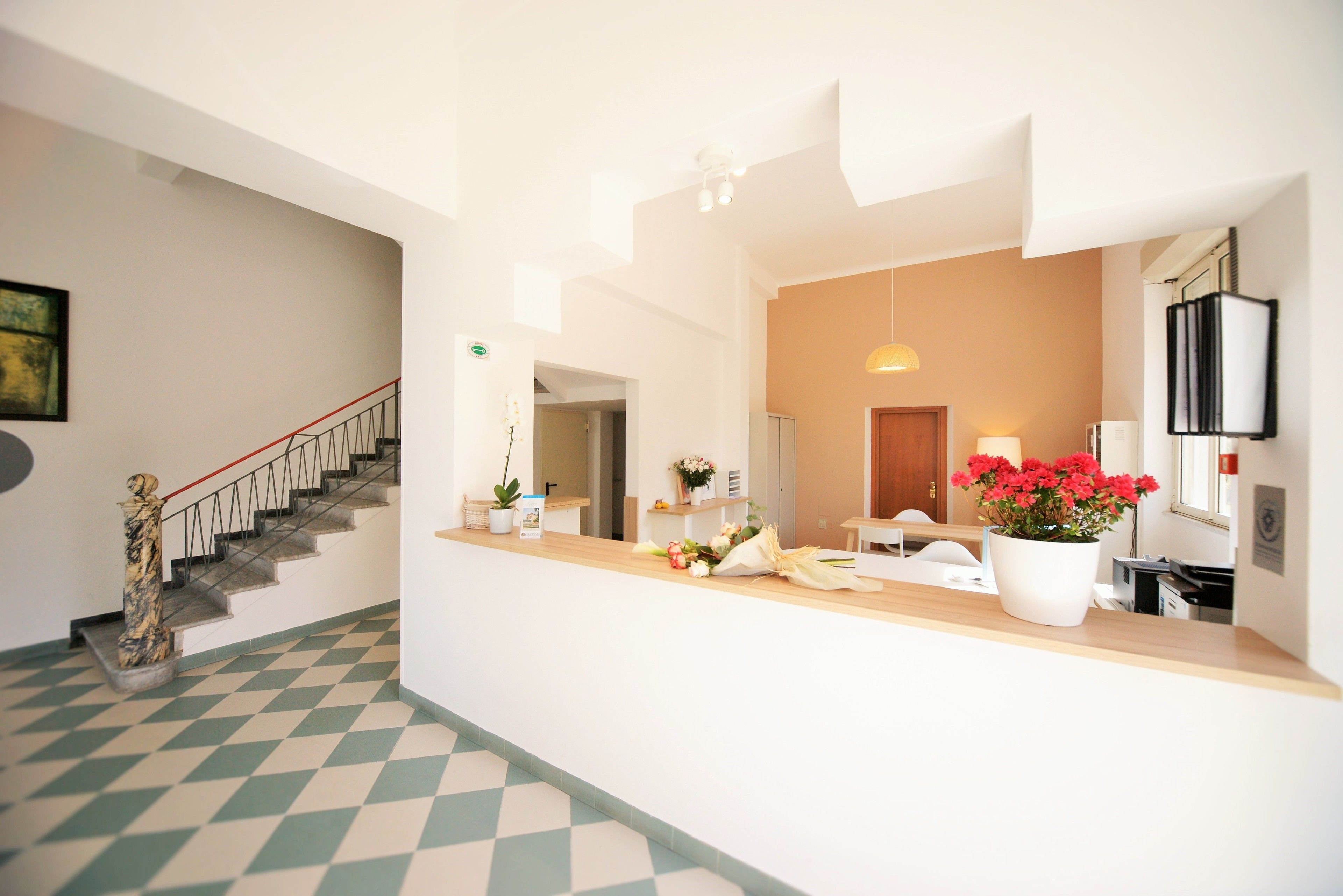 A hotel reception area with a check-in counter, a staircase, and a potted plant with red flowers.