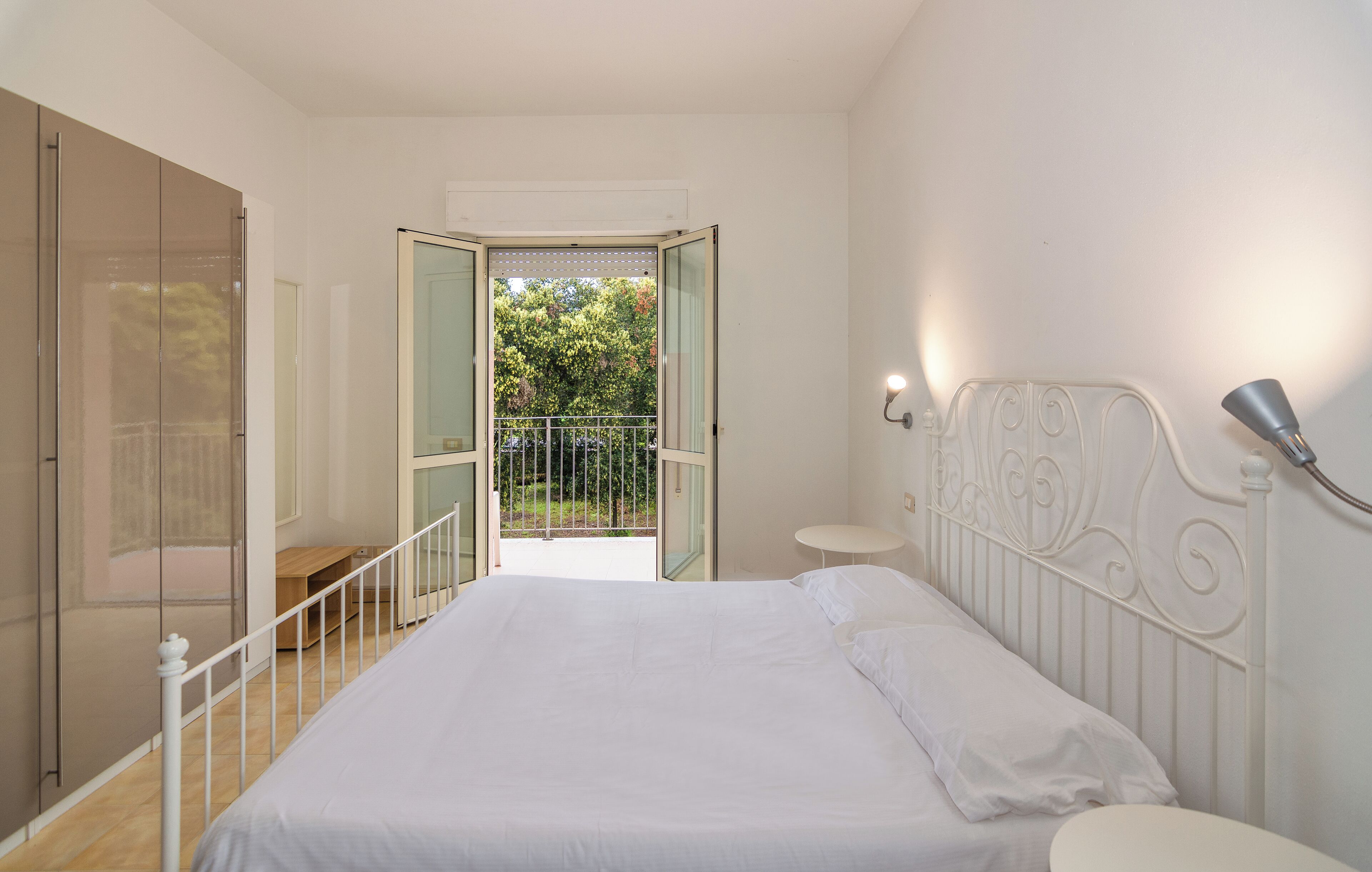 Um quarto com uma cama, uma mesinha de cabeceira, uma pequena mesa, uma varanda com guarda-corpo e uma porta que dá para uma área externa.