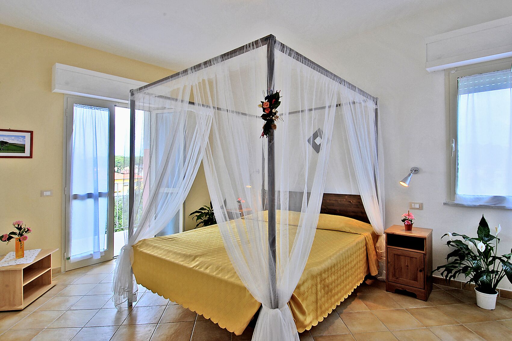 A bedroom with a four-poster bed, a nightstand, a bedside table, and a potted plant.