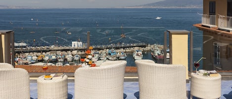 Una terraza en la azotea con mobiliario de mimbre, vista al mar, barcos y una montaña distante.
