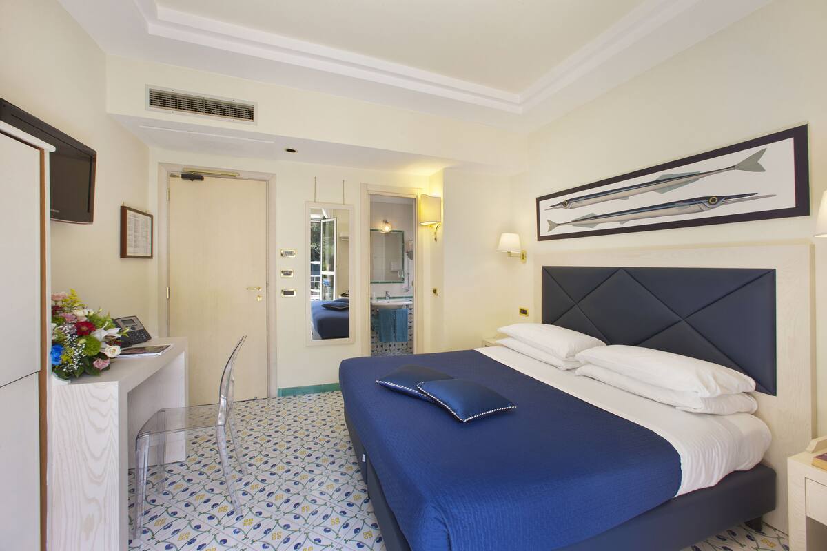 Una habitación de hotel con una cama, un escritorio con teléfono, un espejo y un cuadro en la pared.