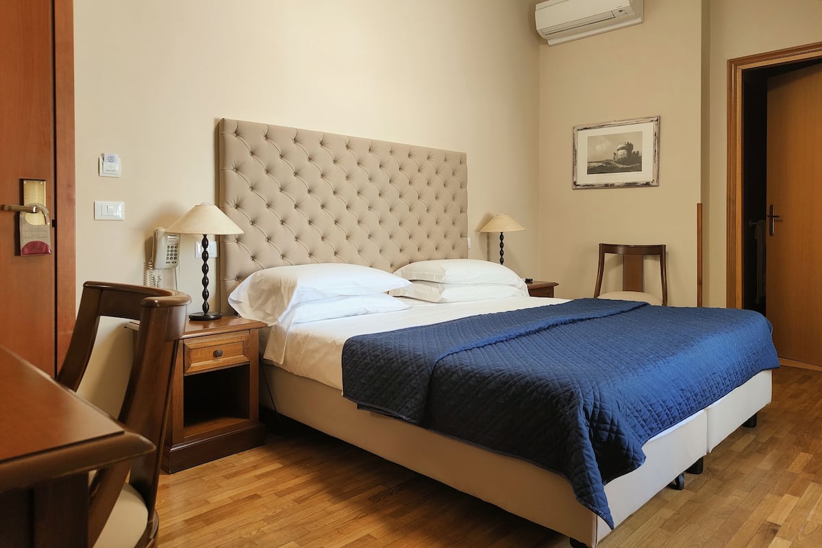 A hotel room with a large bed, two bedside tables with lamps, a wooden chair, a wooden desk, a telephone, an air conditioning unit, and a framed picture on the wall.