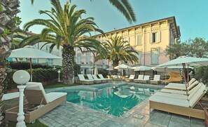 A pool area with lounge chairs and umbrellas, surrounded by palm trees and a building with classical architecture.