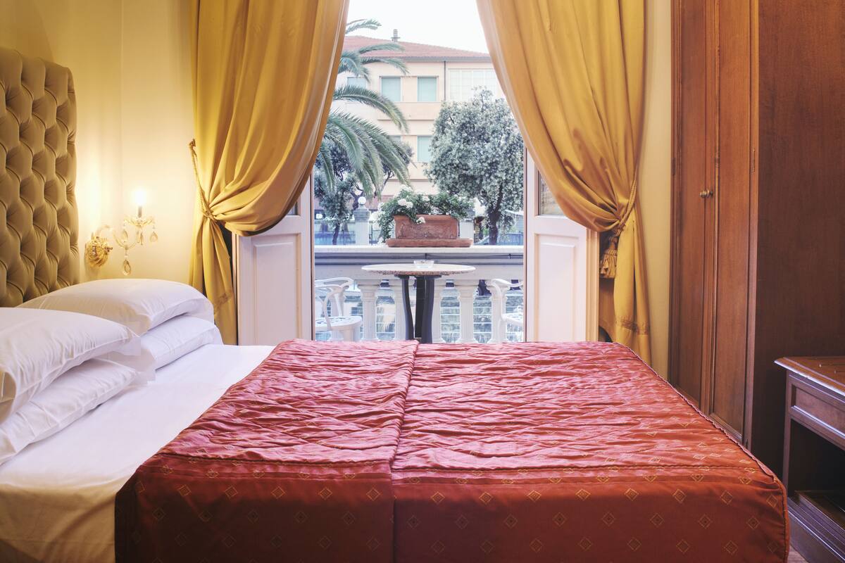 A hotel room with a bed, a window with curtains, a chandelier, and a view of the outside.