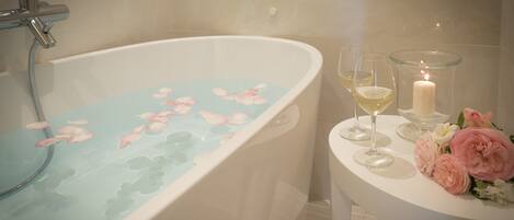 Una bañera con pétalos de rosa, una vela y dos copas de vino blanco sobre una mesa blanca.
