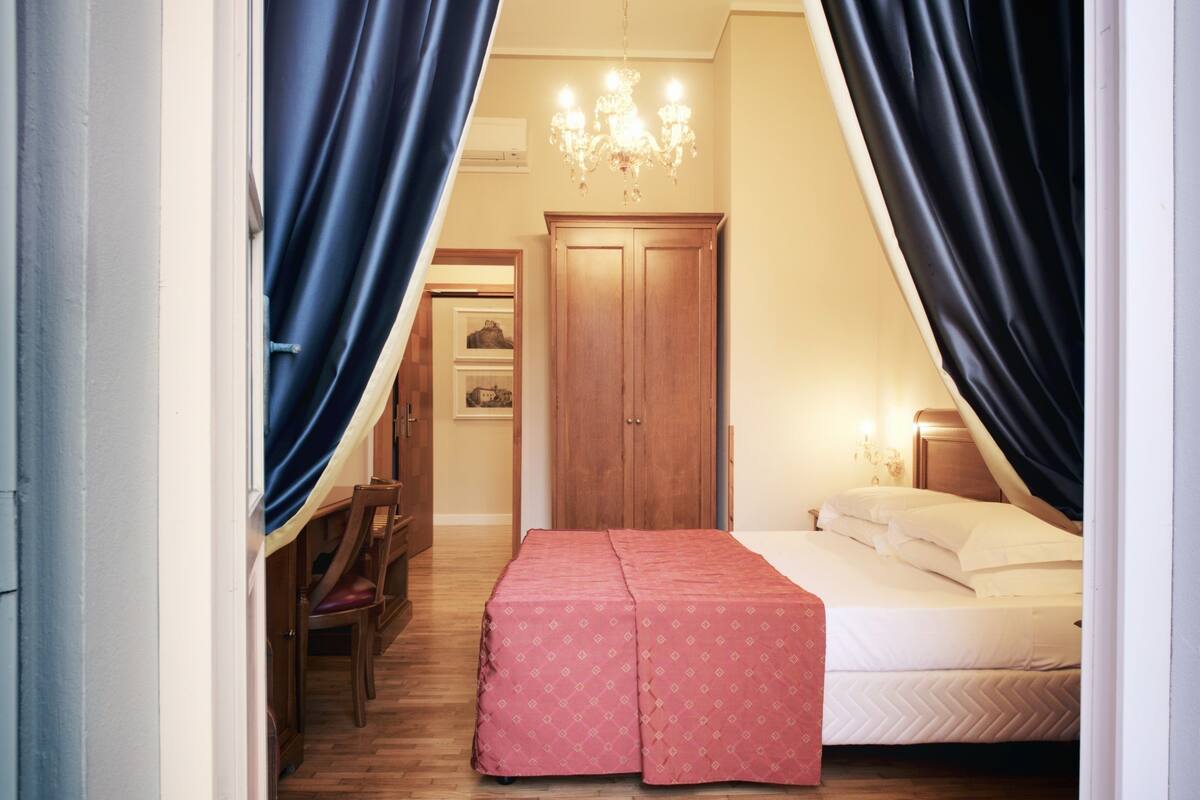 A bedroom with a bed, a wooden wardrobe, a desk with a chair, and a chandelier.