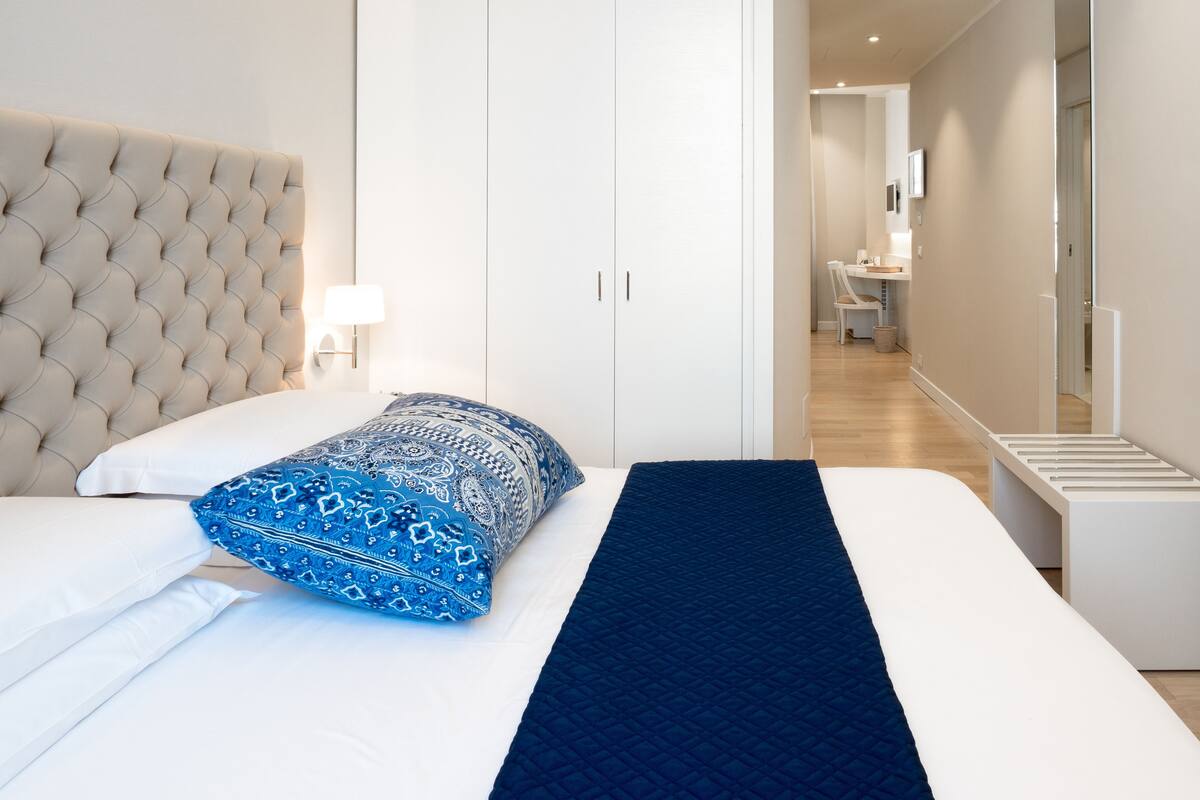 A bedroom with a tufted headboard, white wardrobe, and a desk in the adjacent room.