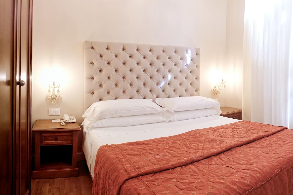 A hotel room with a tufted headboard bed, white linens, a wooden nightstand with a phone, a wooden table with a chair, and a framed picture on the wall.