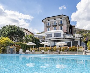 Zona de piscina con sillones reclinables y sombrillas, un edificio grande con balcones y un cielo despejado.