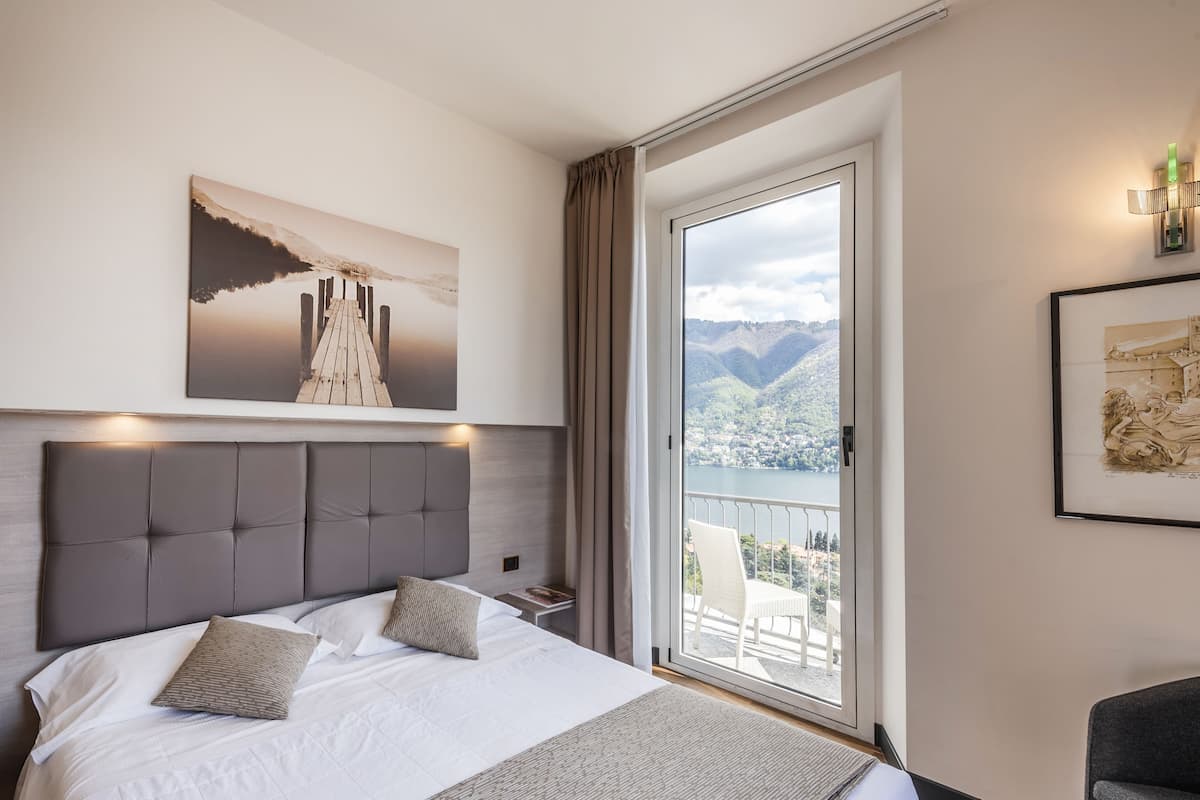 A hotel room with a large bed, a view of a lake and mountains, and a balcony with a chair.