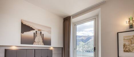 Habitación de hotel con una cama grande, vistas a un lago y montañas, y un balcón con una silla.