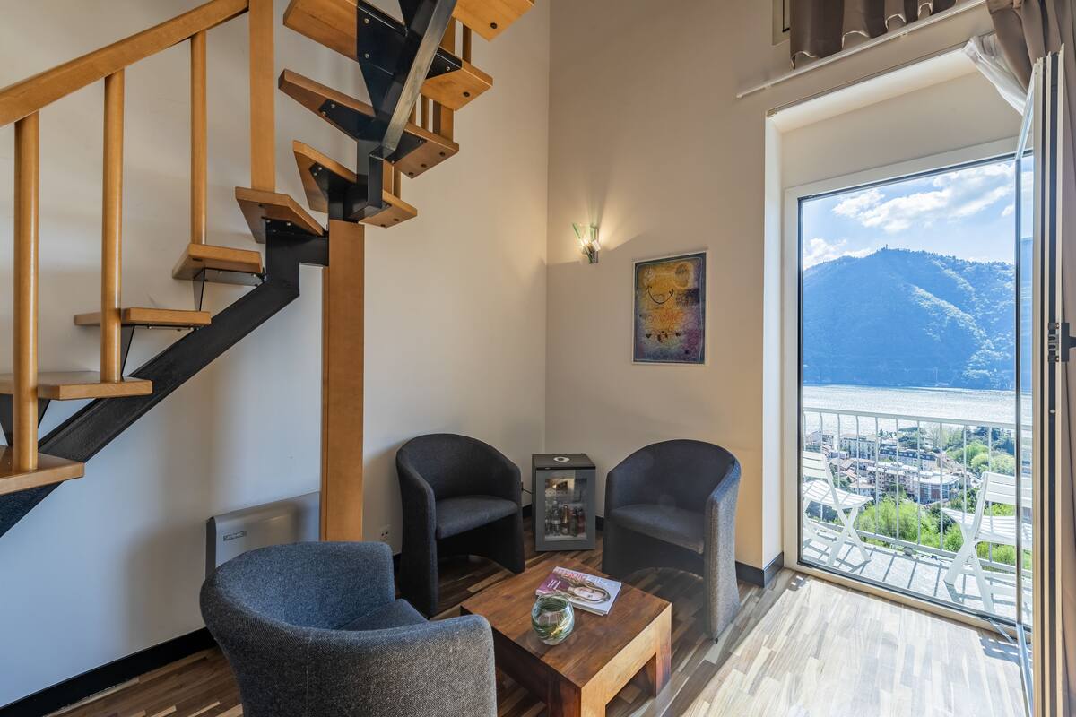 A modern living room with a wooden staircase, two armchairs, a coffee table, and a view of a city and mountains through a large window.