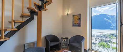 Una sala de estar moderna con una escalera de madera, dos sillones, una mesa de centro y vistas a una ciudad y montañas a través de un amplio ventanal.
