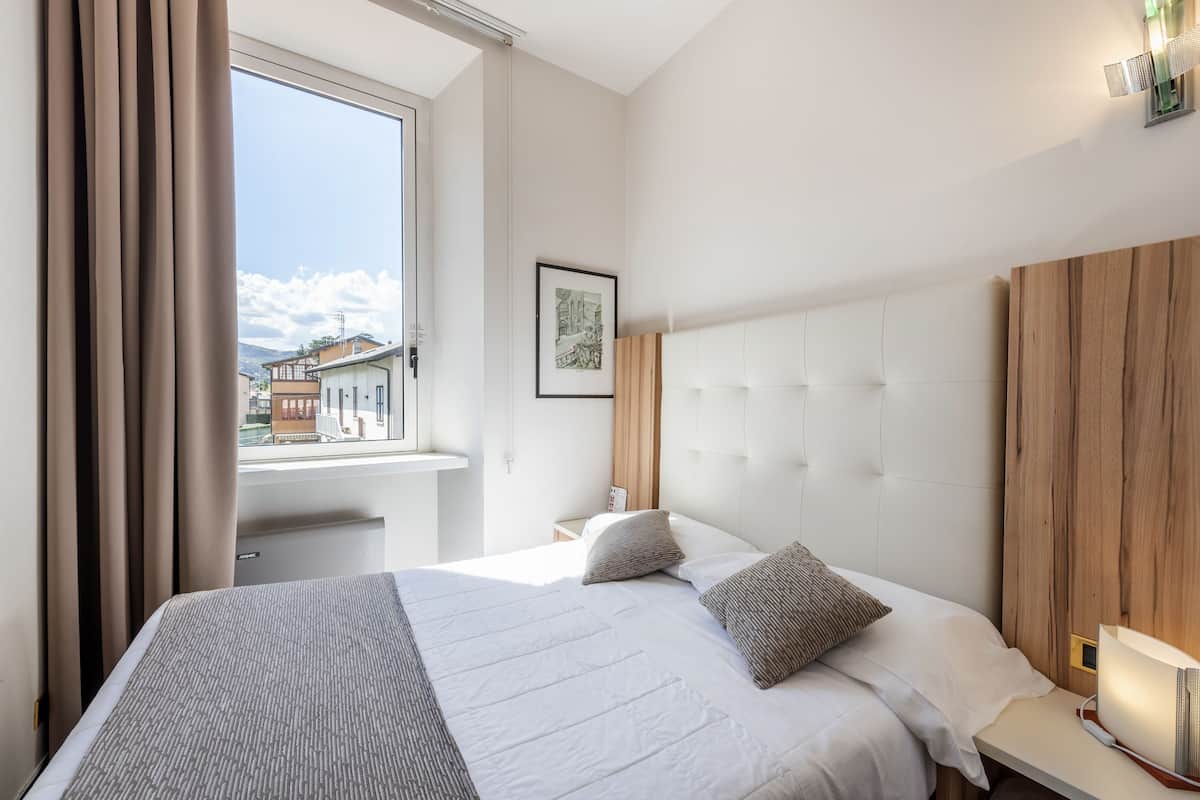 A hotel room with a large bed, a window with curtains, a bedside table with a lamp, and a framed picture on the wall.