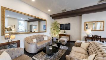 A spacious living room with a large sofa, a coffee table, and a television. There are two large mirrors on the walls, a potted plant, and a small table with a lamp.