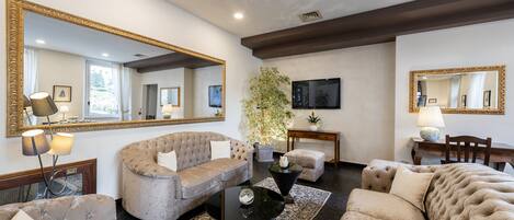 A spacious living room with a large sofa, a coffee table, and a television. There are two large mirrors on the walls, a potted plant, and a small table with a lamp.
