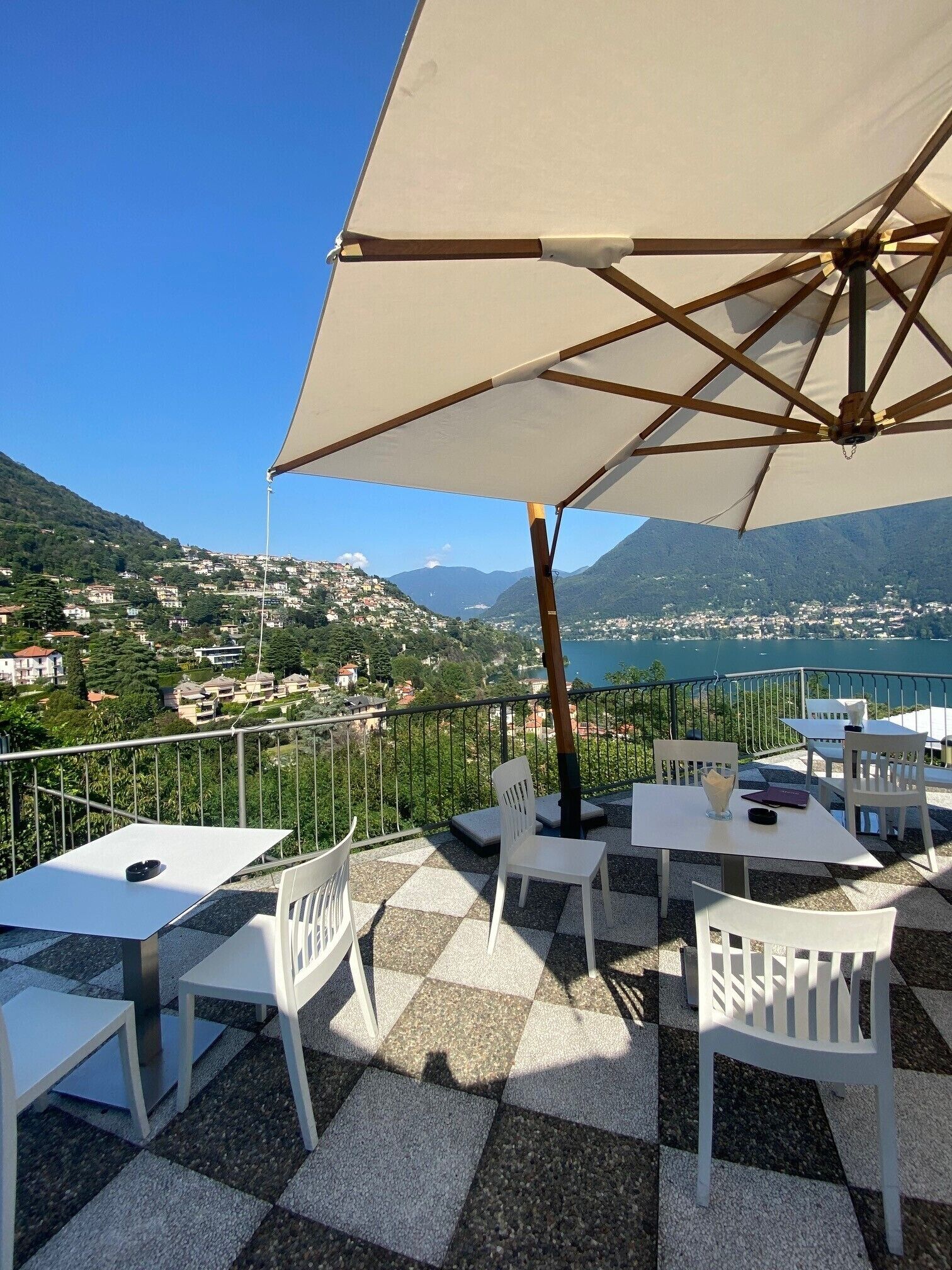 A terrace with checkered flooring, white tables and chairs, and a large umbrella providing shade.