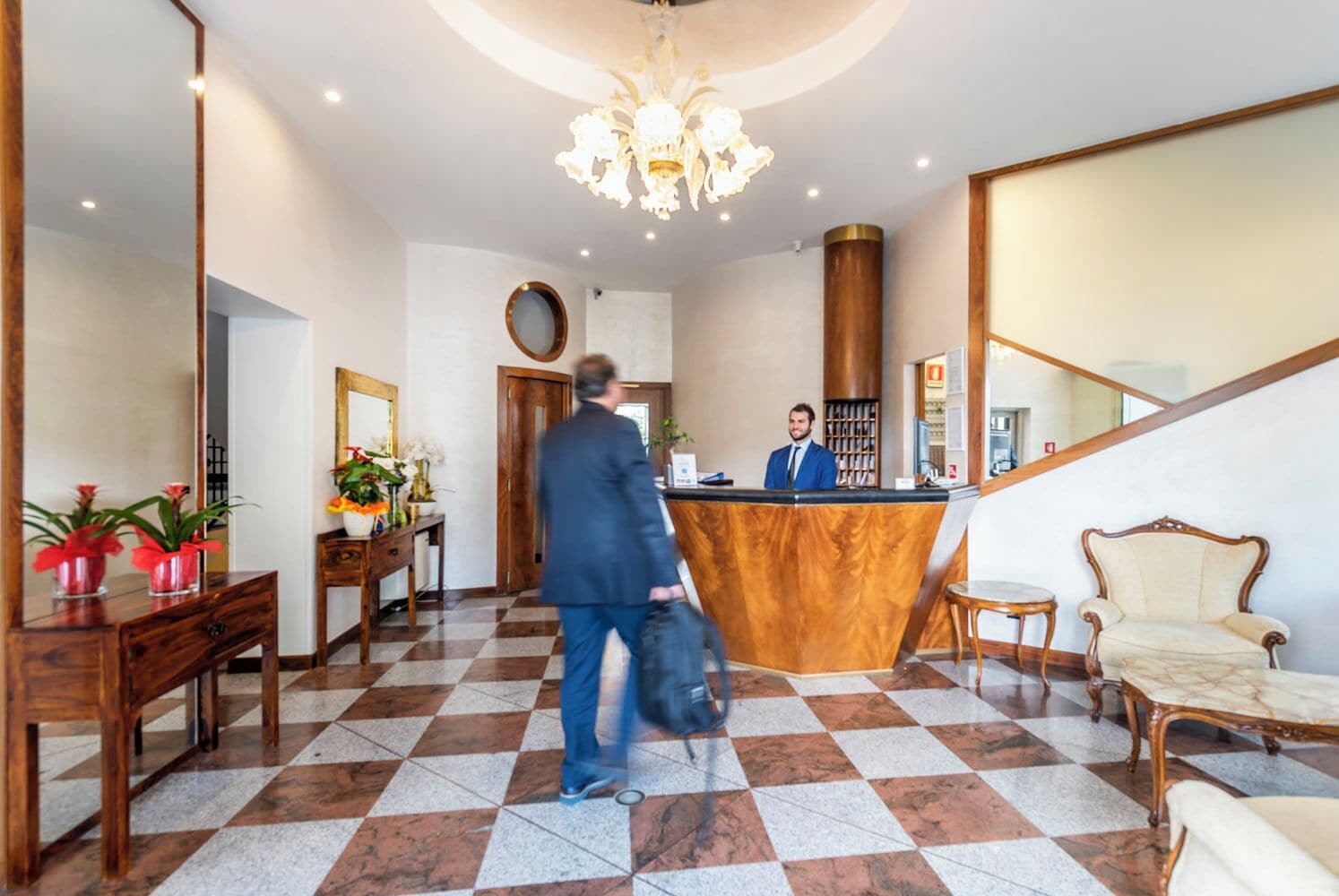 A hotel lobby with a reception desk, a chandelier, a checkered floor, and a large mirror.