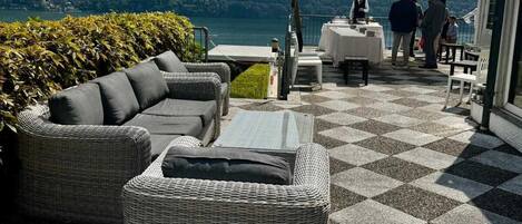 An outdoor seating area with wicker furniture, a checkered floor, and a view of mountains and a lake.
