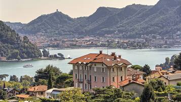 A multi-story building with a red-tiled roof, situated near a body of water with boats, surrounded by greenery and hills.