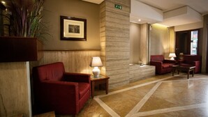 A lobby with a red armchair, a small table with a lamp, a framed picture on the wall, and a pillar with an exit sign.