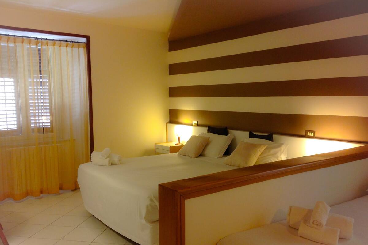 A hotel room with a large bed, bedside lamps, a headboard with horizontal stripes, and a window with curtains.