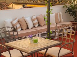Un área de estar en la azotea con sofá, sillas, una mesa con una bebida y una planta en maceta.