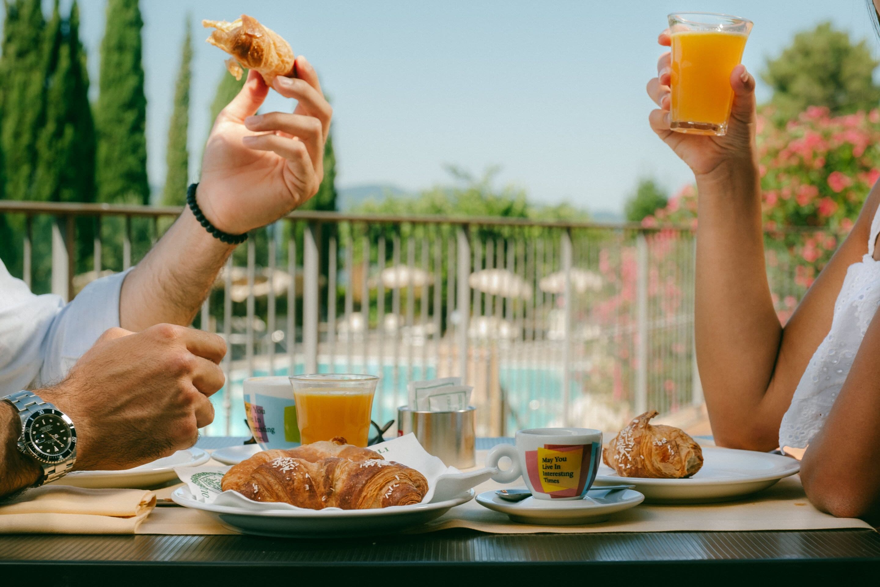 A breakfast setting with croissants, orange juice, and a coffee cup on a table with a view of a pool and greenery.