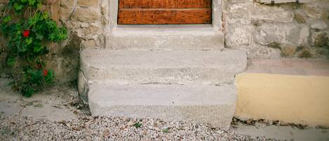 A rustic wooden door with a metal handle, set in a stone wall with a small step leading up to it. There are three signs on the wall indicating different rooms or areas: "Il Giardiniere," "Il Frantoiano," and "Il Cantiniere."