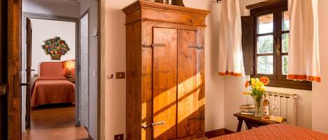 A bedroom with a wooden wardrobe, a bed with a polka dot cover, a window with curtains, and a small table with a vase of flowers.