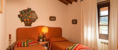 A room with two beds, a wooden ceiling, a window with curtains, and a wall-mounted lamp.