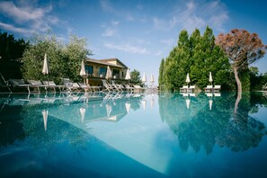 A clear swimming pool with lounge chairs and umbrellas, surrounded by trees and a building with a balcony. - Hotel Fattoria degli Usignoli (Reggello)