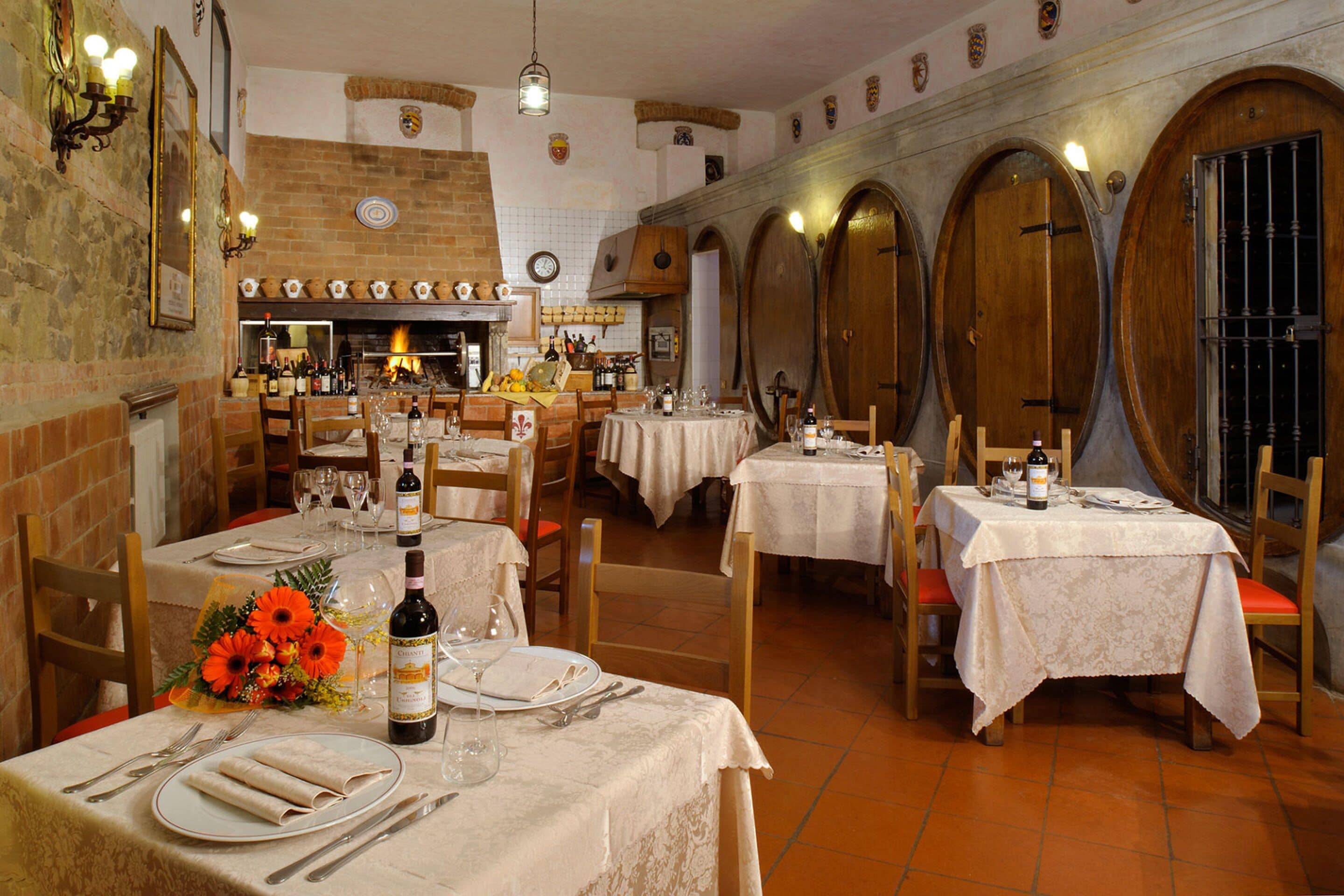 A restaurant with multiple tables set for dining, a brick fireplace, and wooden barrel doors.