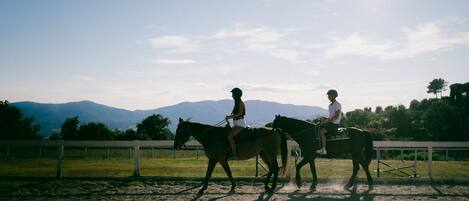 Dos personas montando a caballo en una arena exterior con montañas al fondo.