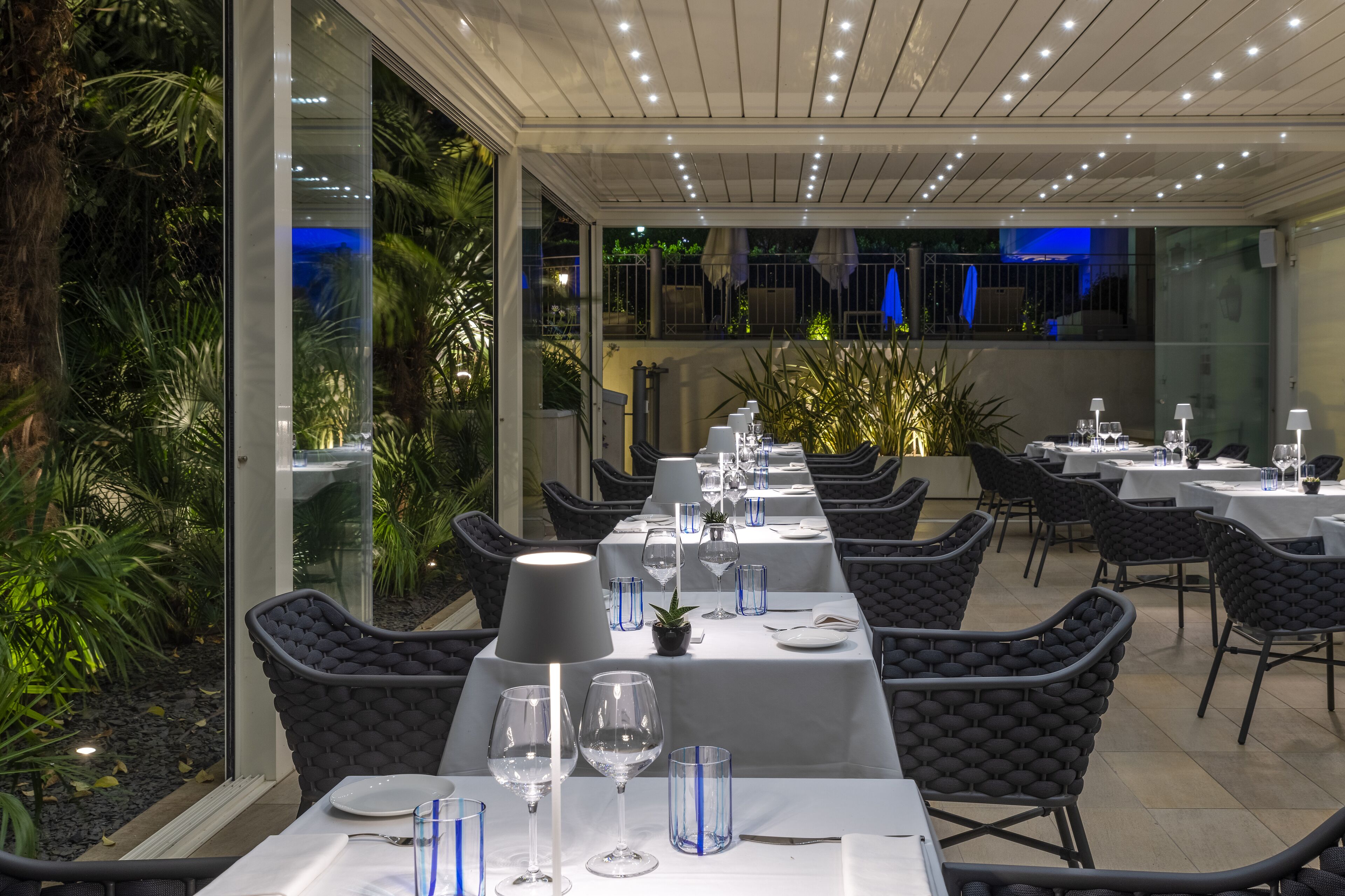 A restaurant with long tables, chairs, and a view of greenery.