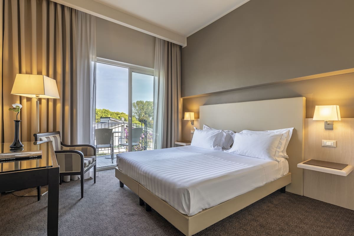 A hotel room with a large bed, two bedside lamps, a desk with a chair, and a view of greenery through the window.