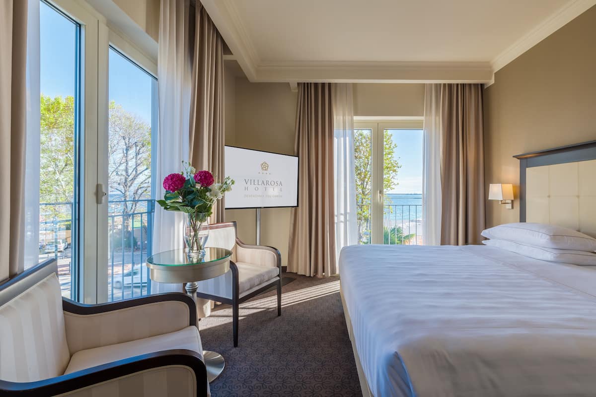 A hotel room with a bed, a chair, a small table with a vase of flowers, a TV, and a balcony with curtains.