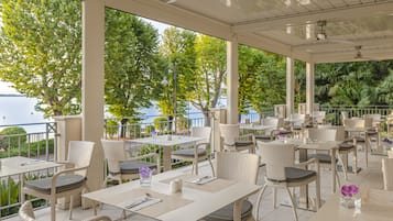 A covered outdoor dining area with tables and chairs, overlooking a body of water and greenery.