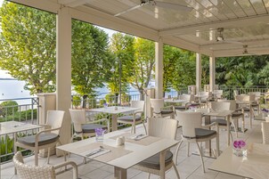 Ein überdachter Essbereich im Freien mit Tischen und Stühlen, mit Blick auf ein Gewässer und Grünflächen.