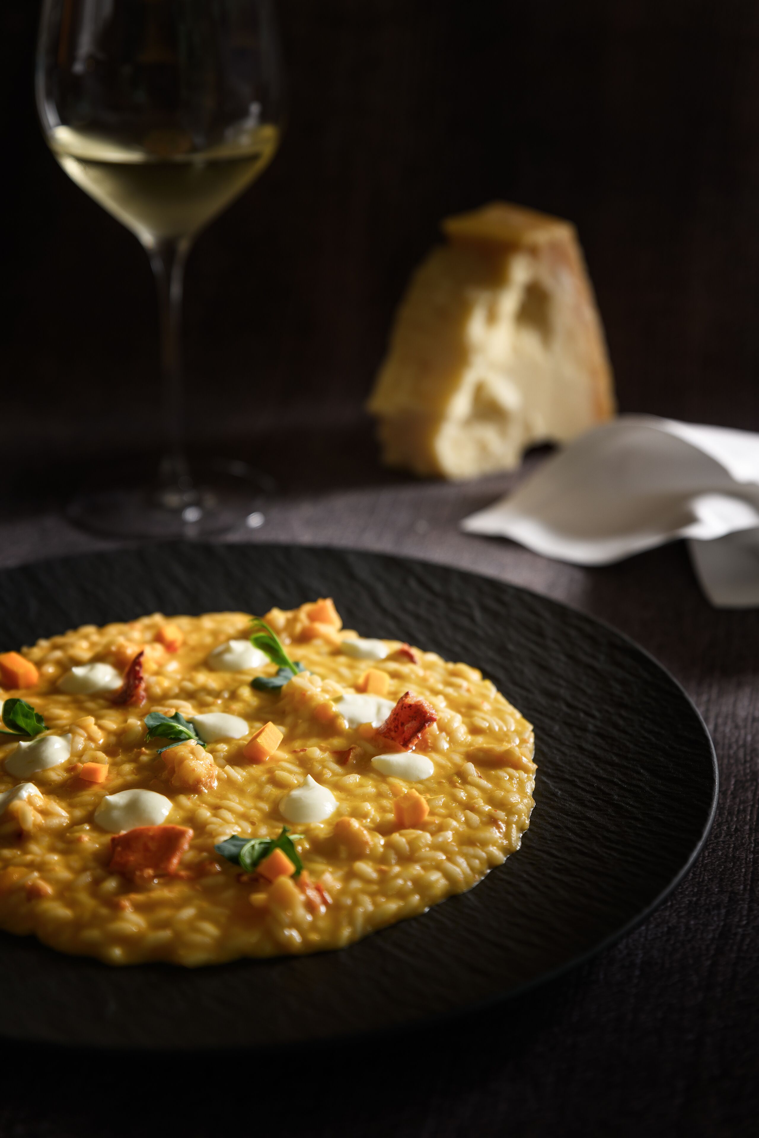 A plate of risotto with cheese and herbs, a glass of white wine, and a loaf of bread on a dark table.