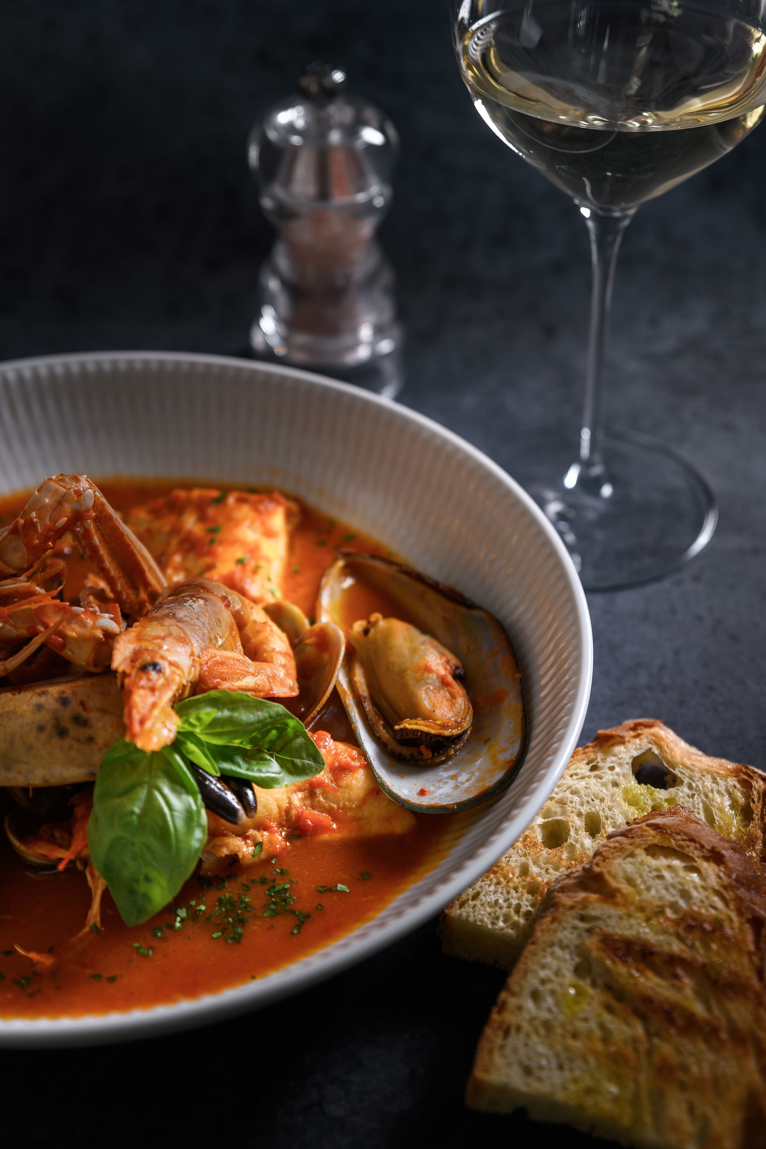 A bowl of seafood stew with bread and a glass of white wine on a dark surface.