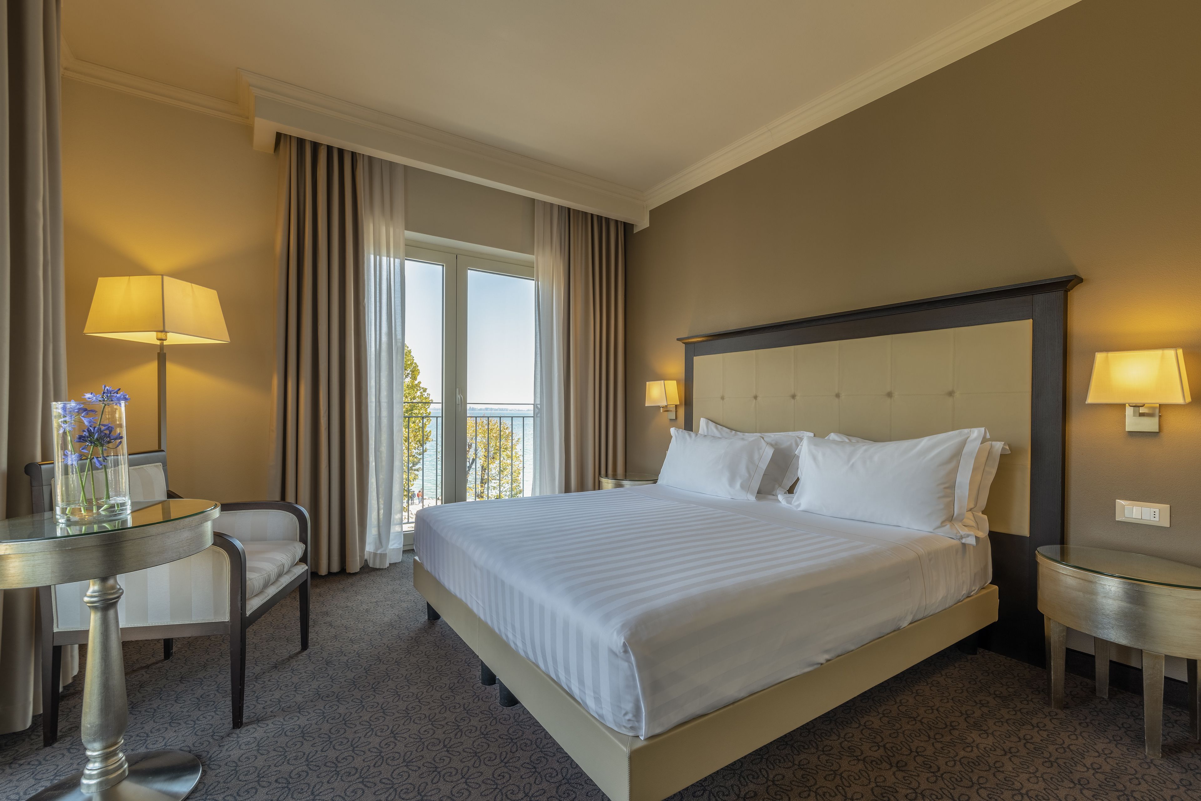 Habitación de hotel con una cama grande, dos mesitas de noche, una silla y un ventanal con cortinas.